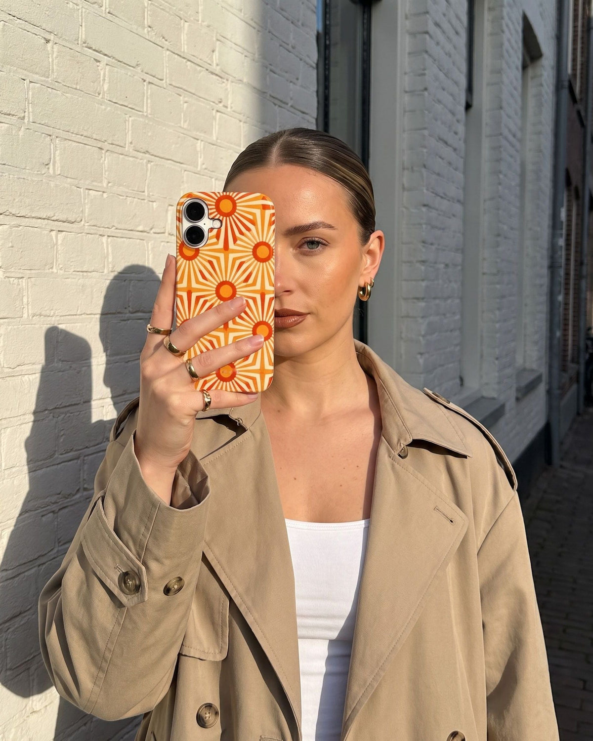 Vrouw in een trenchcoat houdt haar telefoon met het Fynne-hoesje Here Comes Sunny voor haar gezicht, tegen een zonnige witte muur