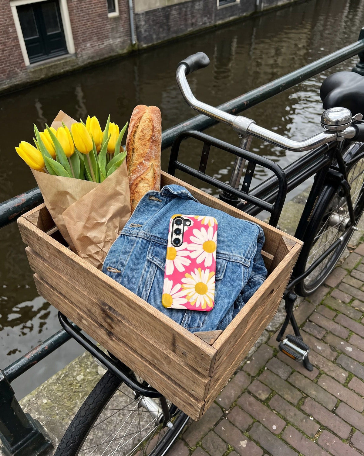 Telefoon met het madeliefjeshoesje Daisy Chain Reaction in een fietskrat met gele tulpen en stokbrood aan de gracht
