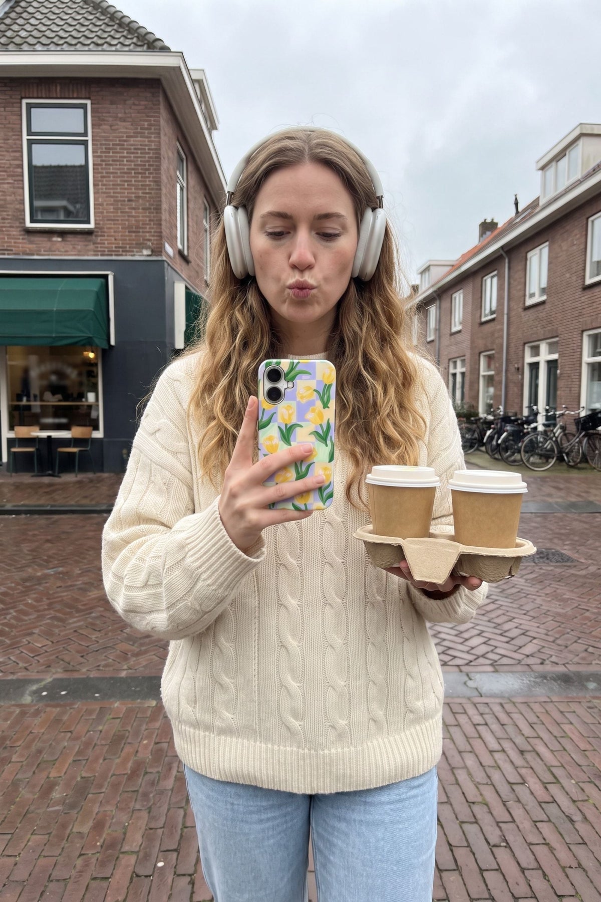 Vrouw met koptelefoon en twee koffie to go kijkt op haar telefoon met het tulpenhoesje Check Mate Tulips