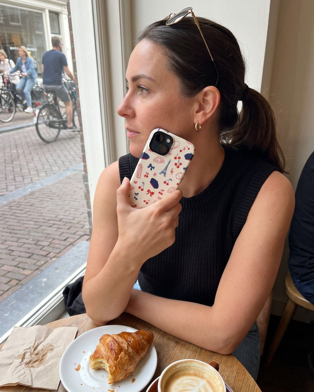 Vrouw bij een caféraam met een croissant houdt haar telefoon met het Fynne-hoesje Très Cherry tegen haar wang
