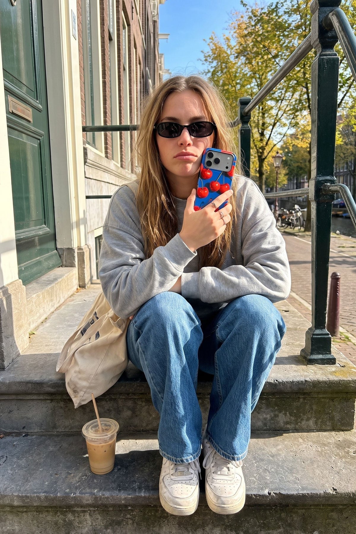 Vrouw met zonnebril op de stoep van een grachtenpand met haar telefoon in het blauwe kersenhoesje Cherry Pop