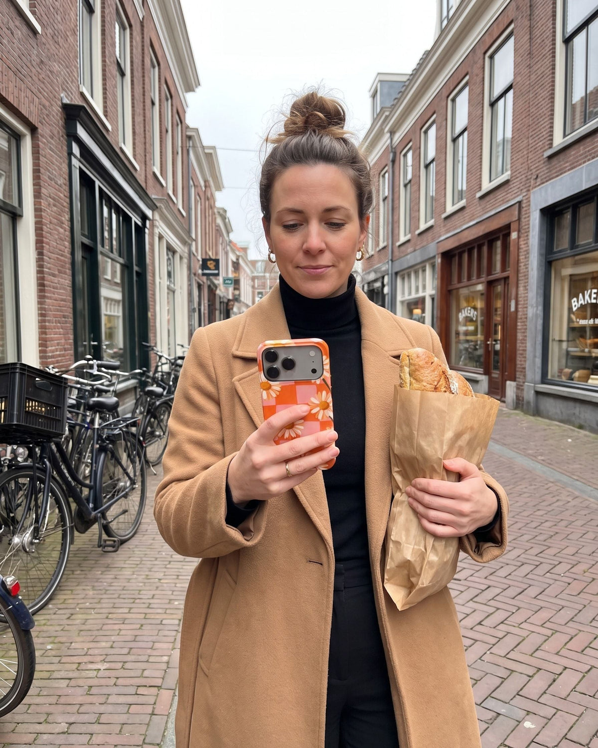 Vrouw met een broodzak in een winkelstraat kijkt op haar telefoon met het Fynne-hoesje Check Your Petals