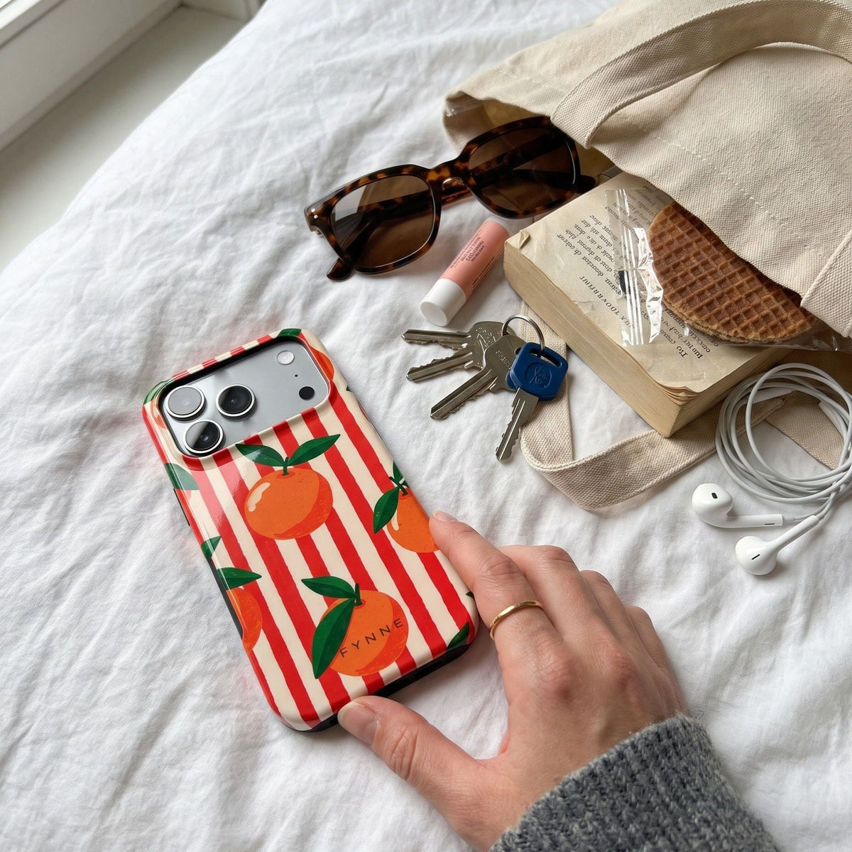 Telefoon met het sinaasappelhoesje Orange You Cute op bed naast een tas, zonnebril, sleutels en een stroopwafel