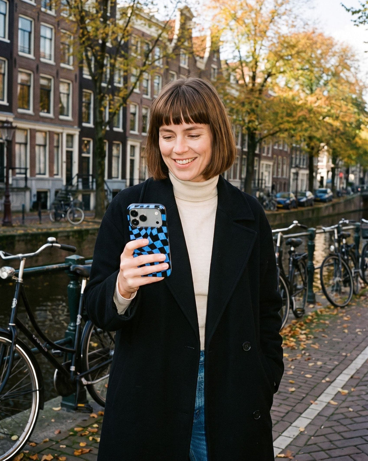 Vrouw aan een Amsterdamse gracht in de herfst kijkt op haar telefoon met het blauwe ruithoesje Check Mate Wobble